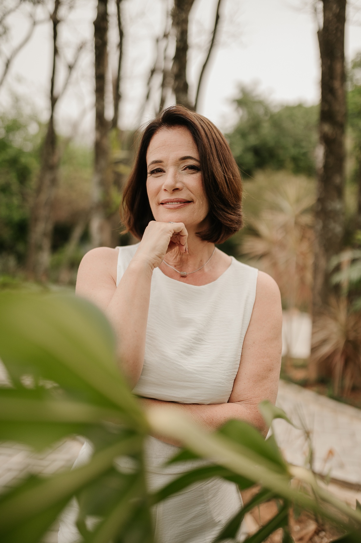 Retrato de mulher madura com vestido branco em ambiente natural, mão apoiada sob o queixo e sorriso sereno entre folhagens