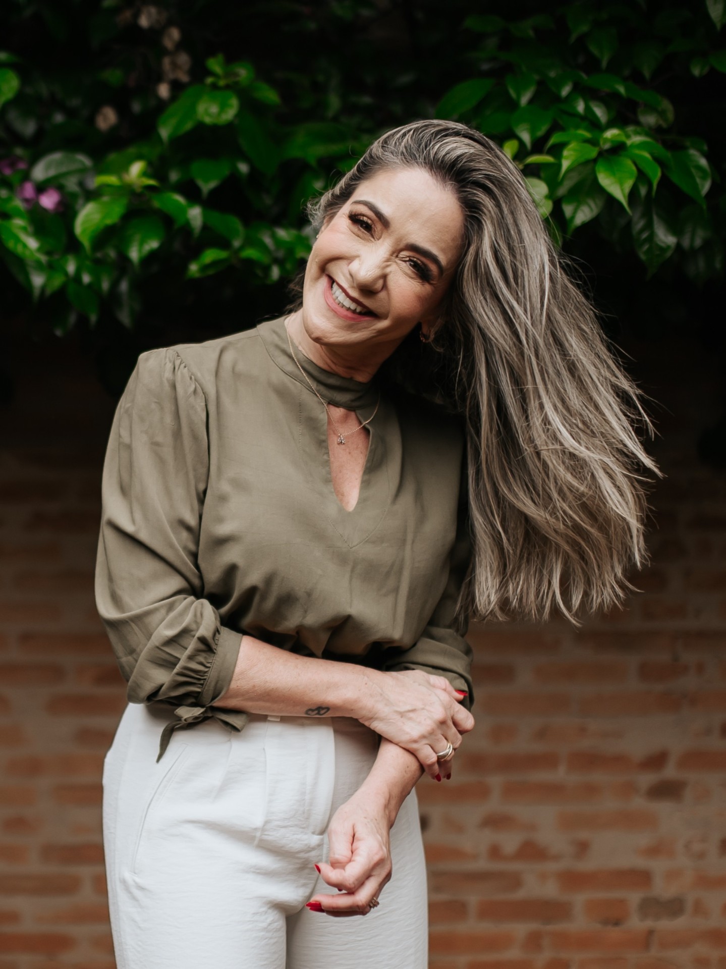 Mulher de cabelos longos grisalhos sorrindo espontaneamente, com blusa verde-oliva, diante de parede de tijolos e folhagem