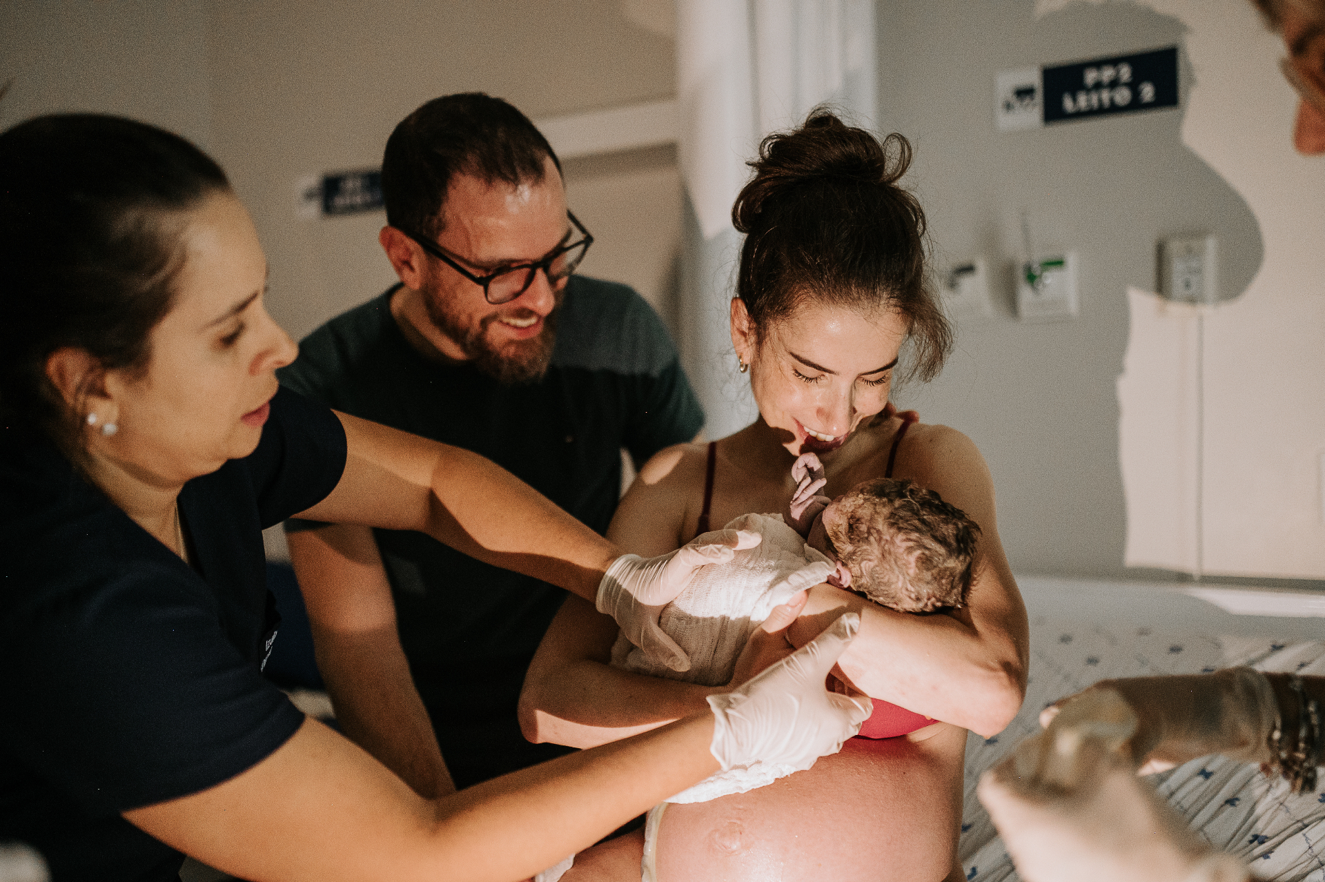 Mãe segurando o bebê recém-nascido logo após o parto, acompanhada pela equipe médica e pelo pai emocionado