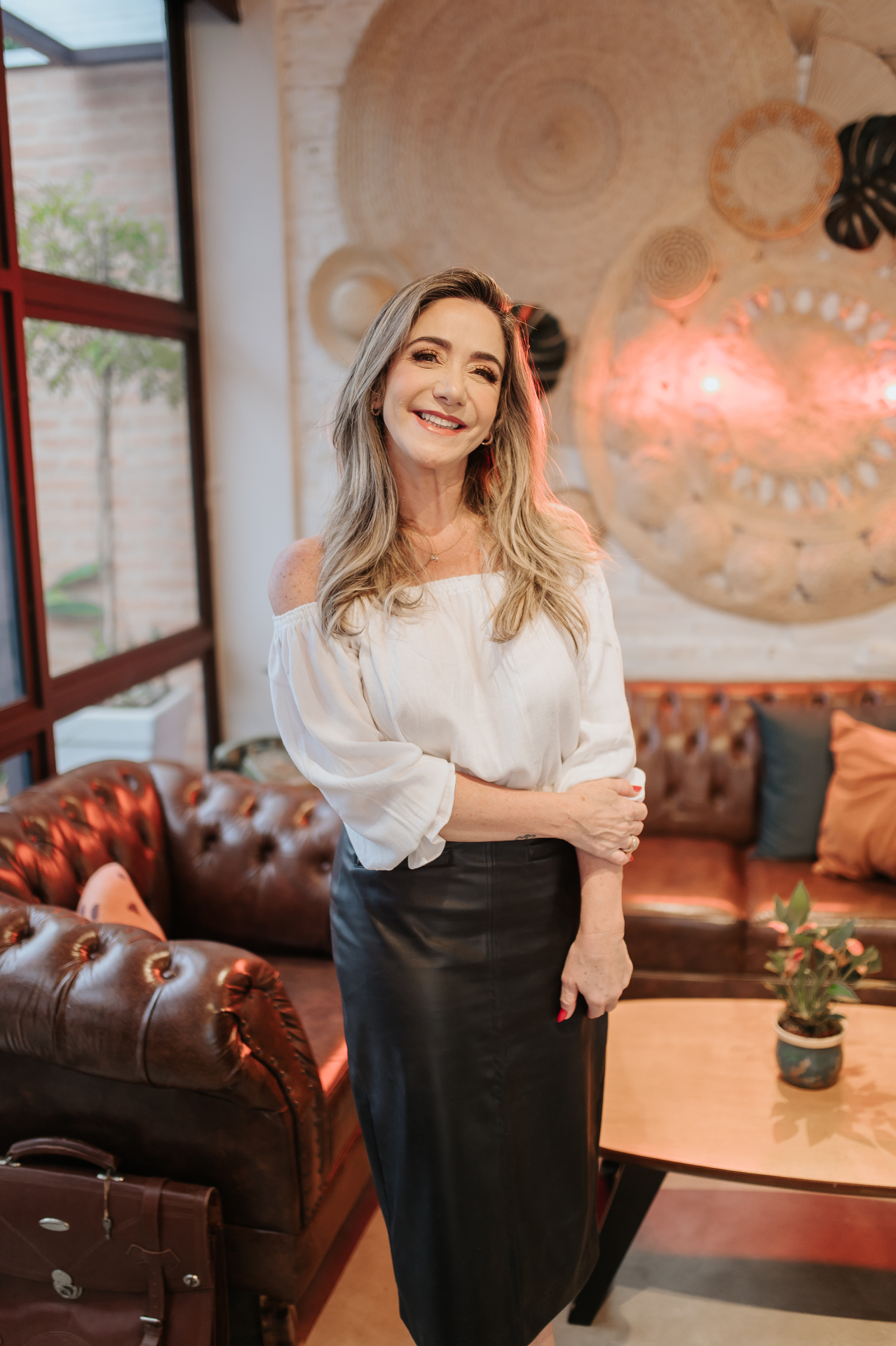 Retrato de mulher com blusa branca e saia preta em pé num lounge com sofás de couro marrom e decoração em fibras naturais
