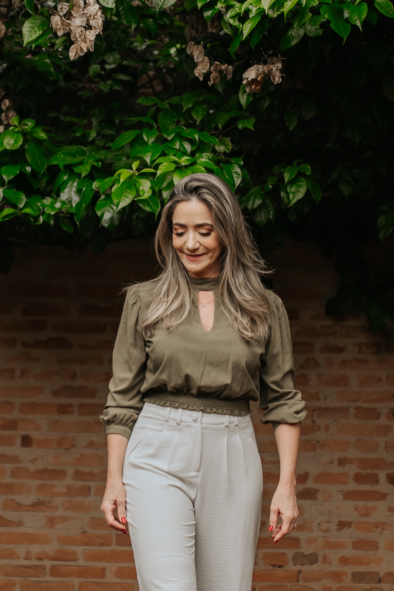 Retrato de mulher com blusa verde-oliva e calça clara diante de parede de tijolos, cabelo ao vento e olhar voltado para baixo em sorriso discreto