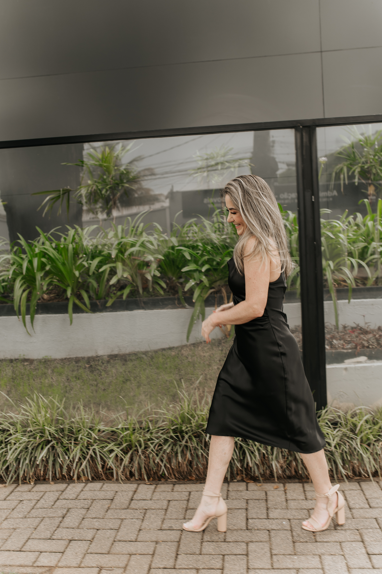 Mulher caminhando de perfil com vestido preto midi e sandálias claras diante de fachada envidraçada com plantas ao fundo