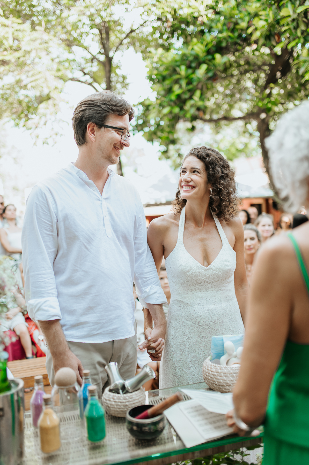 Noivos de mãos dadas, sorrindo um para o outro durante cerimônia intimista ao ar livre sob árvores