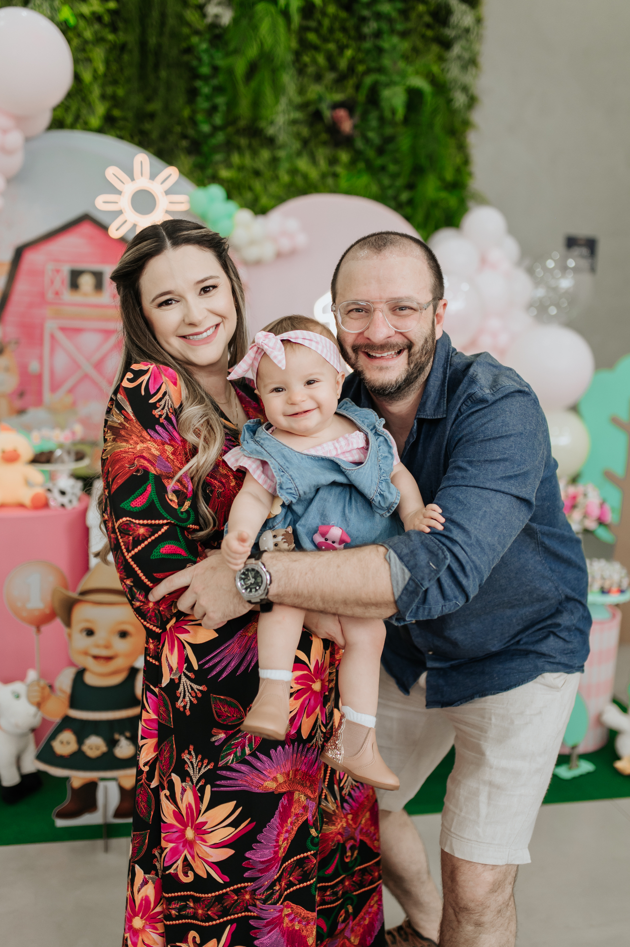 Pais sorridentes segurando a filha bebê em festa de aniversário decorada com balões e cenário temático