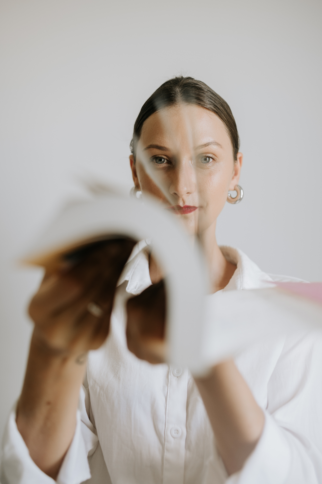 Retrato profissional de mulher com camisa branca, cabelo preso, olhar direto para a câmera com mãos estendidas em desfoque em primeiro plano