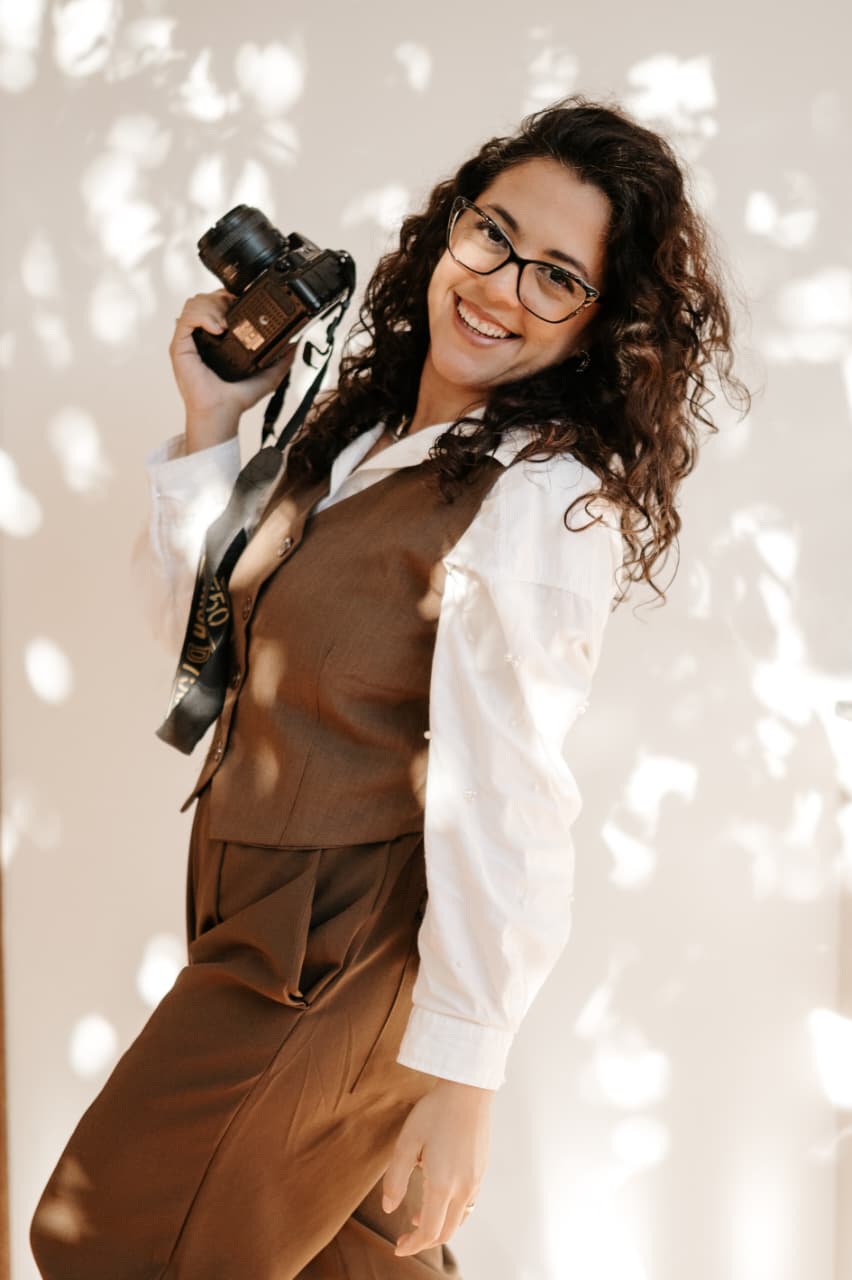 Raquel Lira sorrindo com câmera fotográfica na mão, luz natural filtrada por folhagens