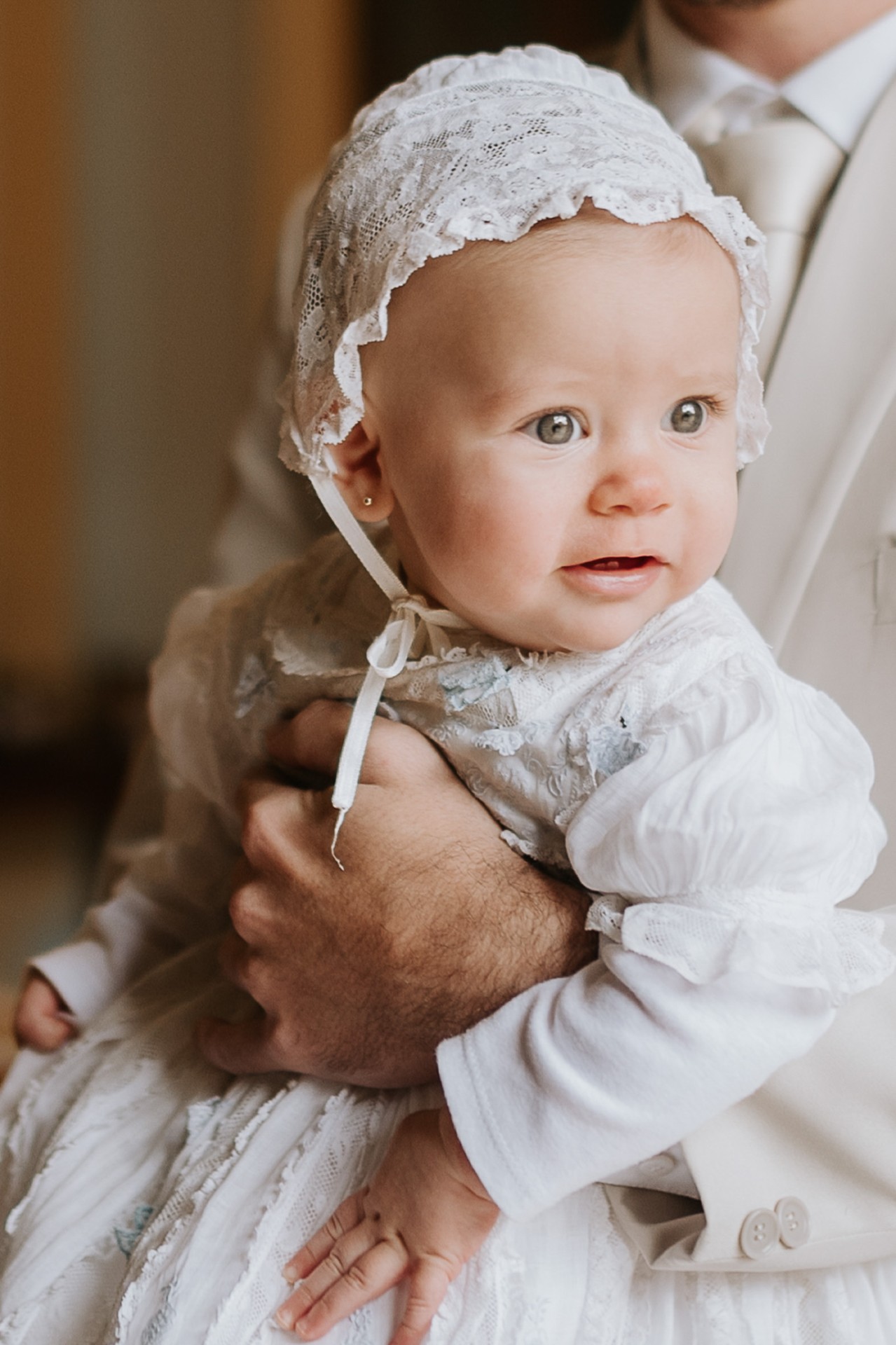 Bebê de olhos claros com touca de renda no colo do pai, vestindo traje branco de batizado