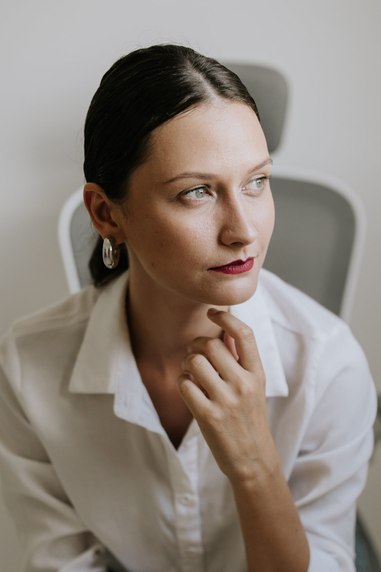 Retrato lateral de mulher com camisa branca, mão apoiada no queixo, olhar contemplativo para o lado em ambiente claro de escritório