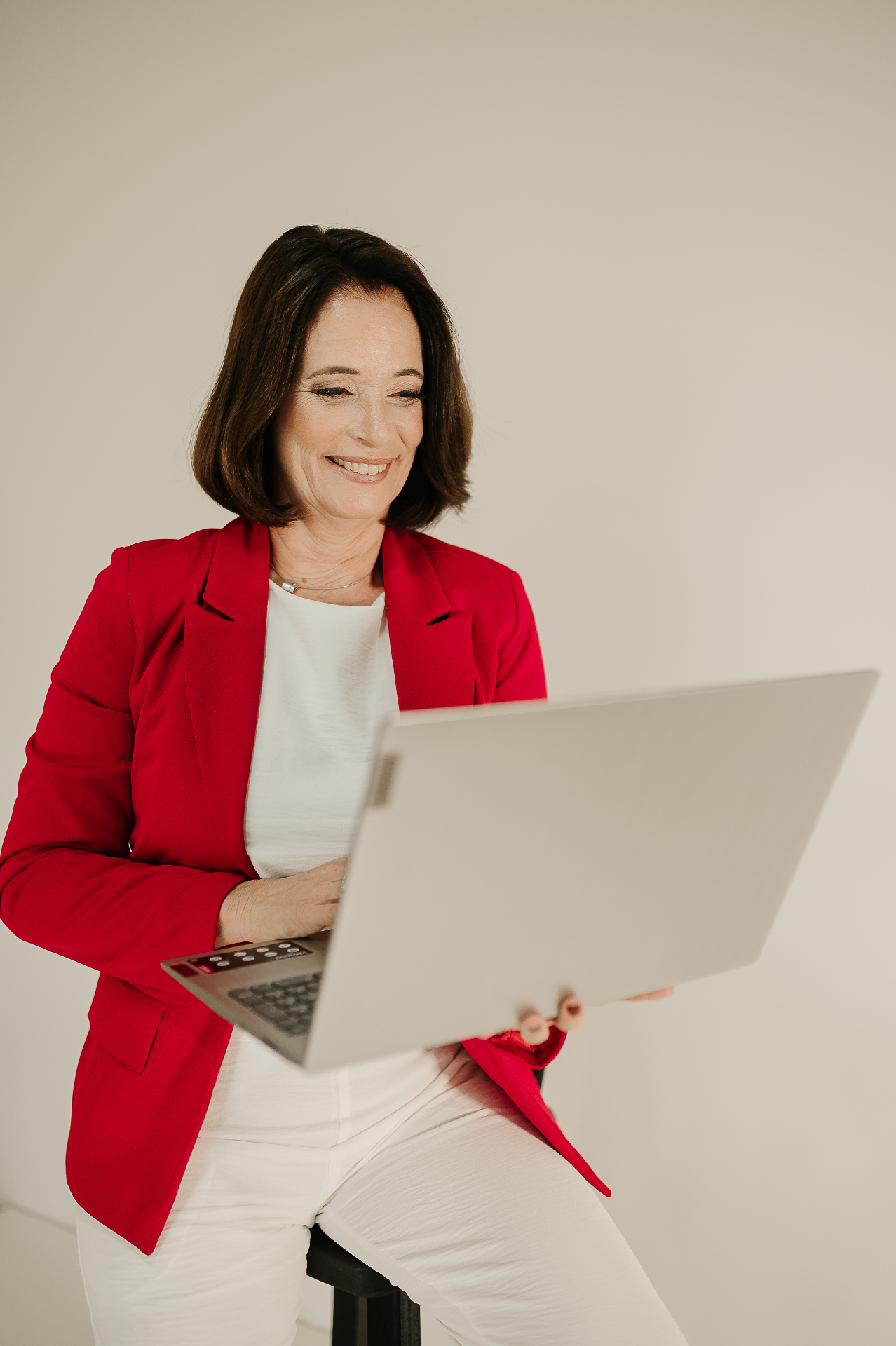 Retrato profissional de mulher madura com blazer vermelho, sentada e sorrindo enquanto trabalha em notebook prata